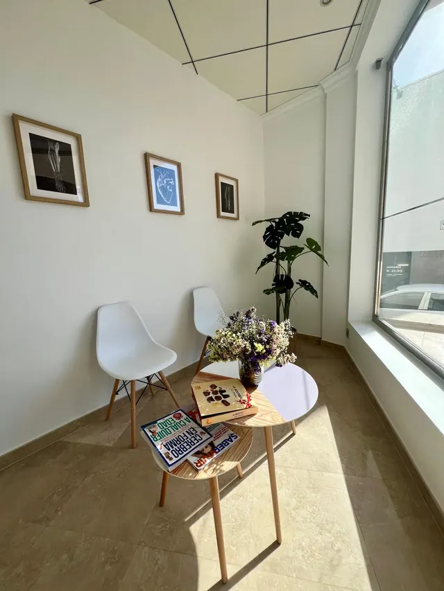 Sala de espera luminosa con sillas blancas, mesas auxiliares de madera, flores y una planta